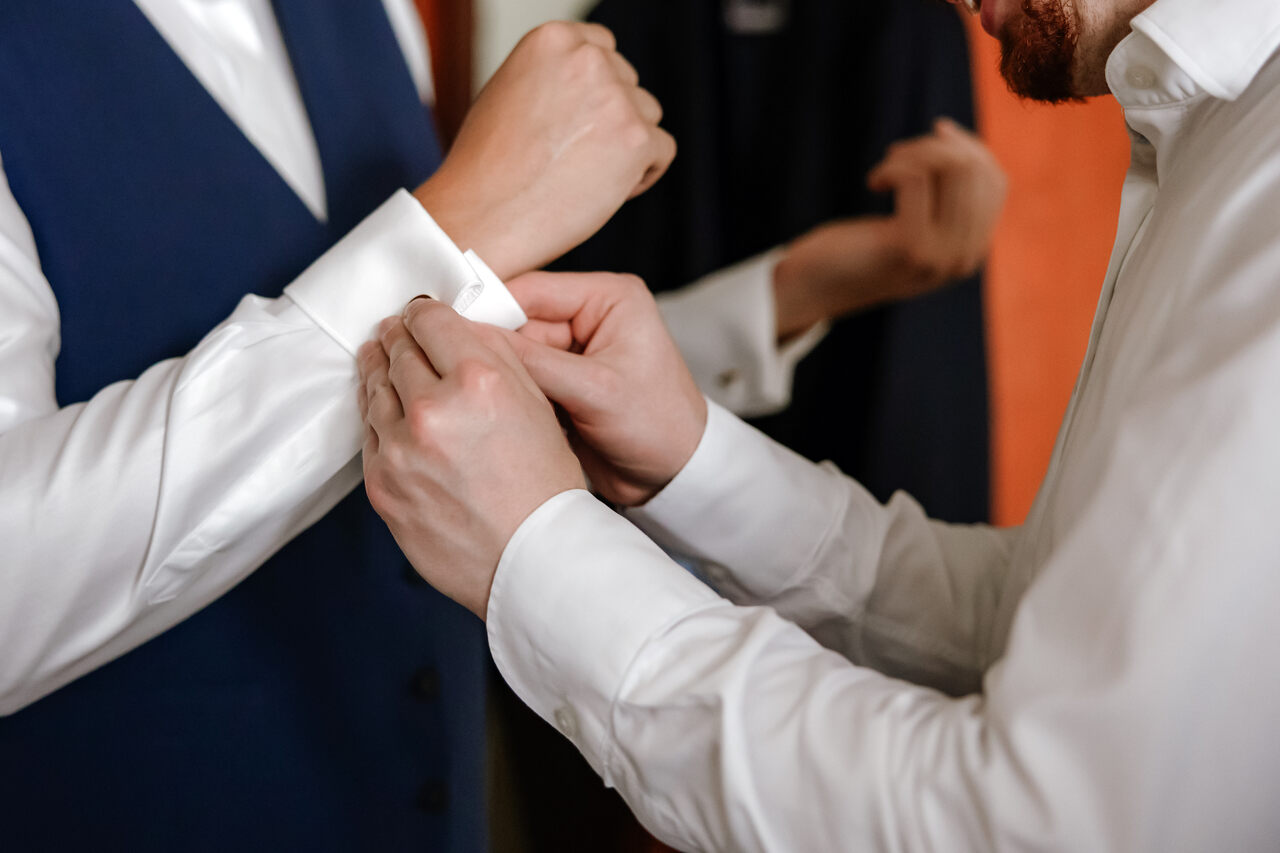 Close nas mãos ajustando o punho de uma camisa social branca, destacando detalhes de alfaiataria masculina.